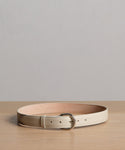 The Classic Belt, crafted from cream nappa leather with a silver buckle, is coiled on a light wooden surface against a plain gray background. This timeless accessory is made in Italy for exceptional style and quality.