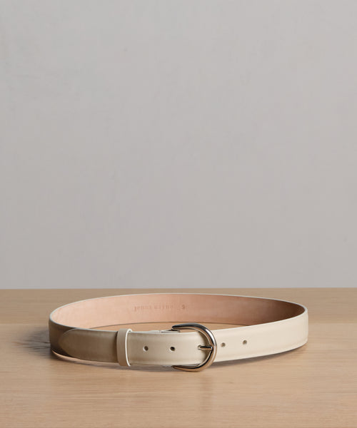 The Classic Belt, crafted from cream nappa leather with a silver buckle, is coiled on a light wooden surface against a plain gray background. This timeless accessory is made in Italy for exceptional style and quality.