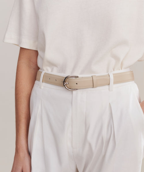 A person wears a short-sleeved white shirt tucked into high-waisted, pleated white pants, styled with the Classic Belt. The look is clean and minimalistic, showing only the torso and upper legs.