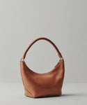 The Crescent Shoulder Bag, crafted from soft brown leather with a rounded base and a single twisted handle, is shown against a plain light gray background.