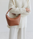 A person in a textured white sweater and cream pants holds the Crescent Shoulder Bag, crafted in Italy from soft leather with a braided handle, against a plain light background.
