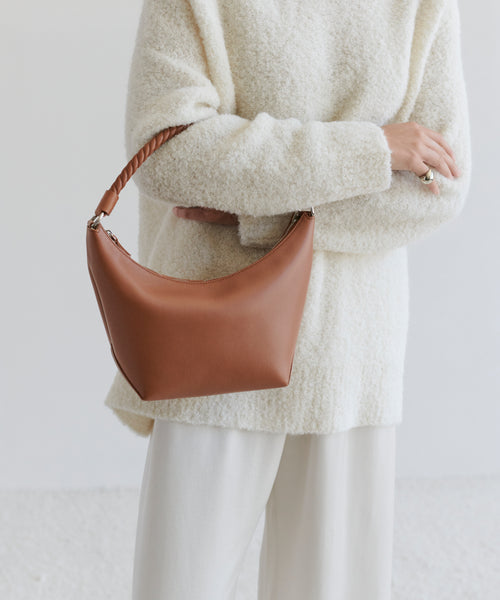 A person in a textured white sweater and cream pants holds the Crescent Shoulder Bag, crafted in Italy from soft leather with a braided handle, against a plain light background.
