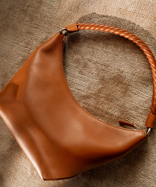 The Crescent Shoulder Bag, handmade in Italy from soft tan leather with a curved silhouette and braided handle, rests on a textured beige surface.