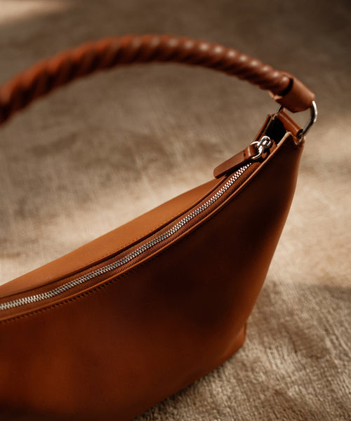 A close-up of the Crescent Shoulder Bag, crafted from soft Italian leather with a twisted thick handle and silver zipper, rests on a textured beige surface under gentle natural light.