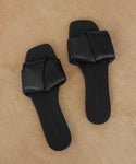 A pair of Dani Slide sandals in black, featuring squared toes and thick padded straps, is displayed on a tan textured surface.