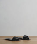 The Dani Slide is a pair of black leather slide sandals featuring wide, knotted straps and a square toe, displayed on a light wooden surface with a plain light gray background.