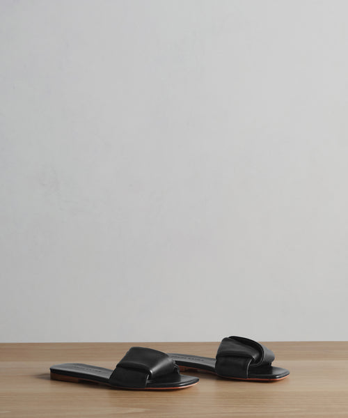 The Dani Slide is a pair of black leather slide sandals featuring wide, knotted straps and a square toe, displayed on a light wooden surface with a plain light gray background.