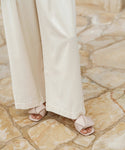 Someone stands on a textured stone floor wearing long, cream-colored pants and the Dani Slide, a light beige leather slide featuring a buttery-soft feel and knot detail.