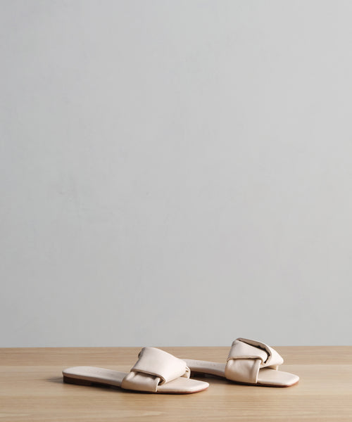 The Dani Slide, a pair of beige buttery-soft leather sandals with padded straps, sits on a light wood surface against a plain gray background.