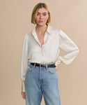 A woman with a blonde bob wears a cream long-sleeve blouse tucked into high-waisted blue jeans, styled with the Leather Danner Belt, standing against a plain beige background for a minimalist look.