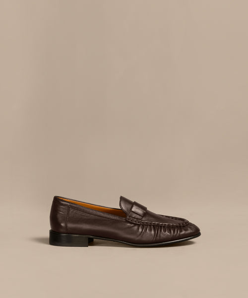 The Leather Dylan Loafer is a dark brown Italian-crafted shoe with a low black heel and moccasin-style stitching, shown in profile against a beige background.