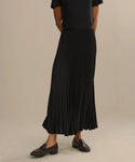 A person wearing a black short-sleeve top, black pleated maxi skirt, and the Leather Dylan Loafer stands against a plain, light background. The persons head is not visible in the image.