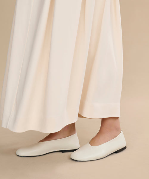 Only the lower legs and feet are visible as someone stands on a beige floor, wearing a long cream-colored skirt paired with Leather Elodie Flats.