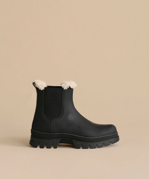 The Leather Highland Boot is a black water-resistant Chelsea style with chunky ridged soles, elastic side panels, and white shearling trim—perfect for stylish winters. Shown against a beige background.