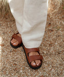 Wearing tan linen pants and Hutton Slide Sandal, a person stands on a gravel path with grass in the background.