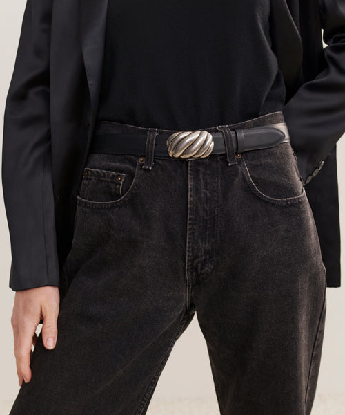 A person wearing high-waisted black jeans with a tucked-in black top, a black blazer, and the Leather Jules Belt featuring a silver swirl rope-twist buckle, with one hand in their pocket.