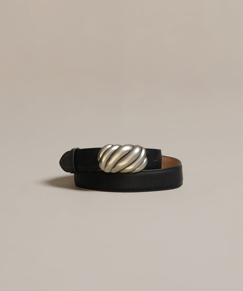 The Leather Jules Belt, a black leather belt with a large rounded rope-twist vintage silver buckle, is coiled and displayed on a neutral beige background.