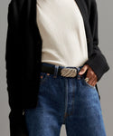 A person wears a black cardigan, white shirt, blue jeans, and the Leather Jules Belt with a rope-twist buckle; one hand is tucked in a pocket.