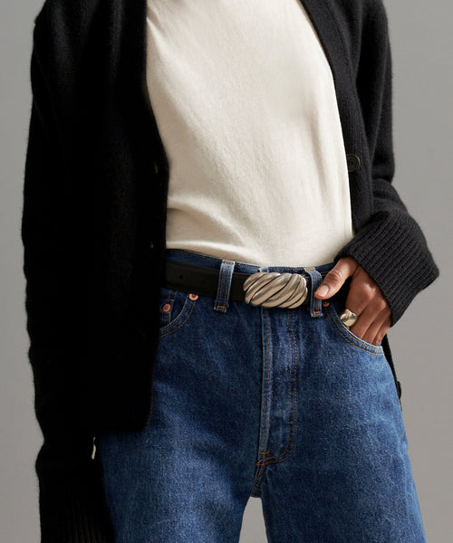 A person wears a black cardigan, white shirt, blue jeans, and the Leather Jules Belt with a rope-twist buckle; one hand is tucked in a pocket.