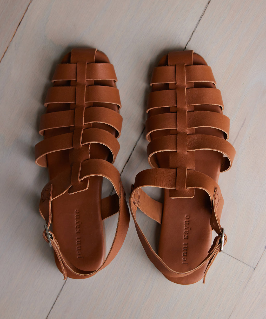 The Leather Lake Sandal, a handmade brown leather fisherman sandal with ankle straps and buckles from Portugal, is displayed side by side on a light wooden floor.