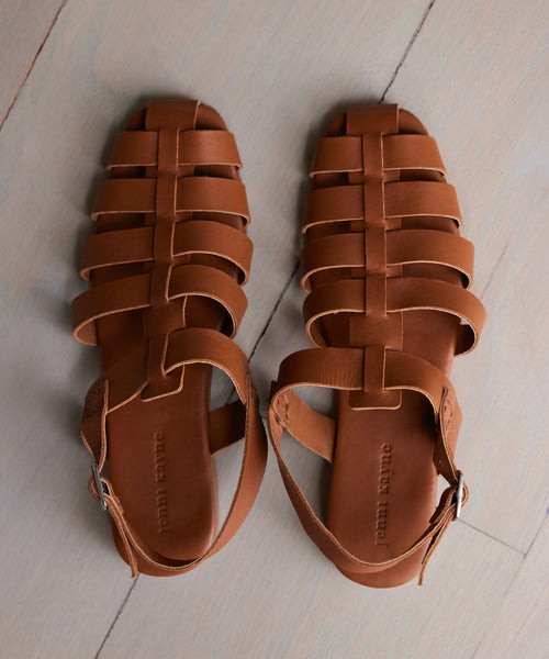 The Leather Lake Sandal, a handmade brown leather fisherman sandal with ankle straps and buckles from Portugal, is displayed side by side on a light wooden floor.