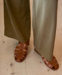 A person wearing olive green wide-leg pants and Leather Lake Sandals, handcrafted in brown woven leather in Portugal, stands on a light wooden floor.