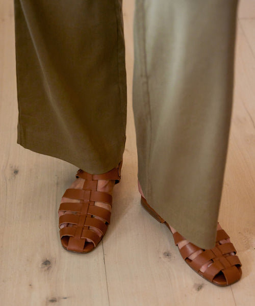 A person wearing olive green wide-leg pants and Leather Lake Sandals, handcrafted in brown woven leather in Portugal, stands on a light wooden floor.