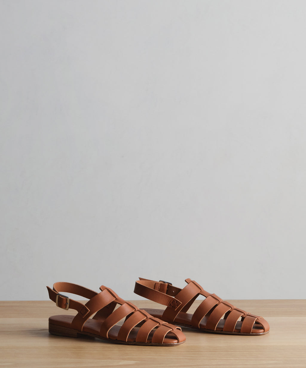 The Leather Lake Sandal, a pair of brown open-toe fisherman sandals with woven straps, handmade in Portugal, is displayed on a wooden surface against a plain light gray background.