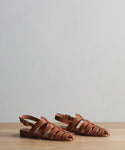 The Leather Lake Sandal, a pair of brown open-toe fisherman sandals with woven straps, handmade in Portugal, is displayed on a wooden surface against a plain light gray background.