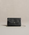 The Loren Clutch Bag is a black leather clutch featuring a silver button clasp and thin strap, crafted in Italy and shown on a neutral beige background.