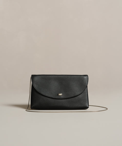 The Loren Clutch Bag is a black leather clutch featuring a silver button clasp and thin strap, crafted in Italy and shown on a neutral beige background.