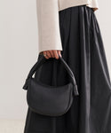A person in a light beige long-sleeve top and black skirt holds the Loren Shoulder Bag by its handles at their side. The Loren features soft pebbled leather and boasts a simple, elegant design.