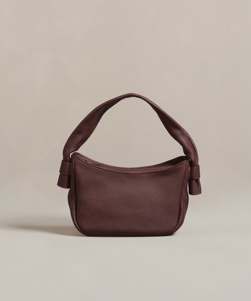 The Loren Shoulder Bag, a small Italian-made accessory, features dark brown soft leather, a curved handle, and knot details on each side, shown against a plain beige background.
