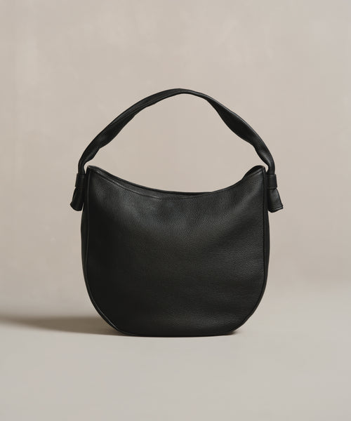 A black Loren Tote, crafted in Italy with a single wide strap and gently rounded, minimal design, is shown against a neutral beige background.