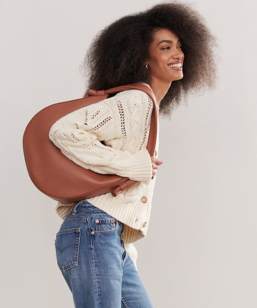 A woman with curly hair smiles, wearing a cream knit sweater and blue jeans, carrying the Loren Tote—a spacious leather bag—over her shoulder as she stands sideways against a light background.