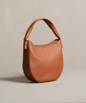 The Loren Tote is a tan leather bag with a rounded bottom, wide shoulder strap, and zipper closure, expertly crafted in Italy and shown against a plain light beige background.