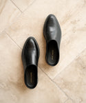 A pair of Leather Lug Mules in black leather with rounded toes, shown on a light beige tiled floor. The Joan Tayre brand name is visible inside each shoe.
