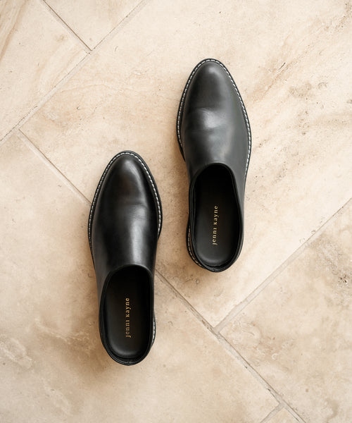 A pair of Leather Lug Mules in black leather with rounded toes, shown on a light beige tiled floor. The Joan Tayre brand name is visible inside each shoe.