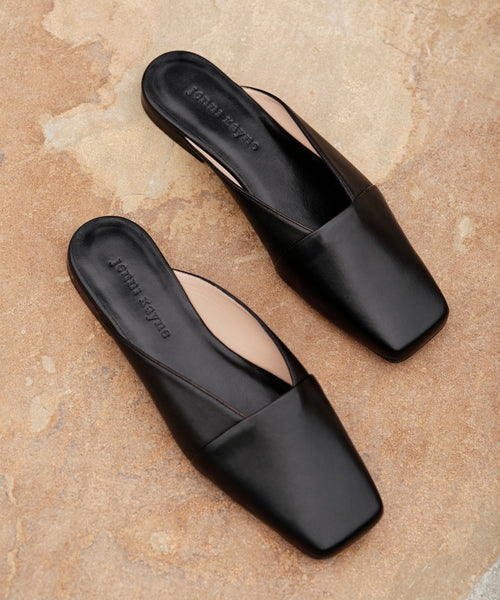A pair of Mia Mule black, square-toe Italian leather mules with John Wayne embossed on the insole, displayed on a textured light brown surface.