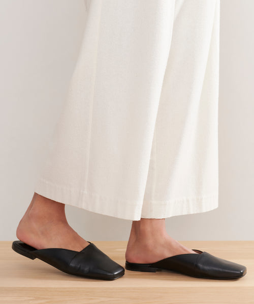 Person wearing loose, white, ankle-length pants and Mia Mule black Italian leather square-toe flats, standing on a light wooden floor against a plain background.