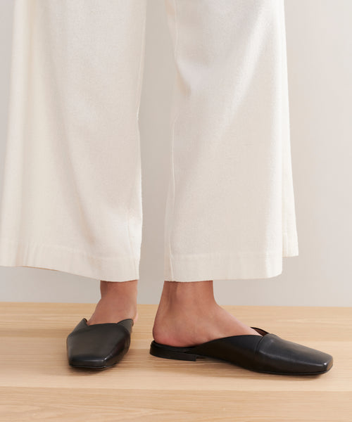 A person wears white wide-leg pants and black Mia Mules with a square toe, standing on a light wood floor against a neutral background.