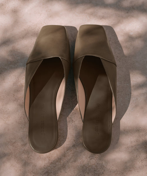 A pair of Mia Mule brown square-toe slip-on shoes is placed side by side on a light, textured surface with dappled sunlight and soft shadows.