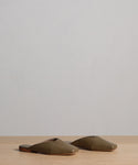 The Mia Mule, a pair of olive green, closed-toe Italian leather mules, rests on a light wooden surface with a plain light gray background.