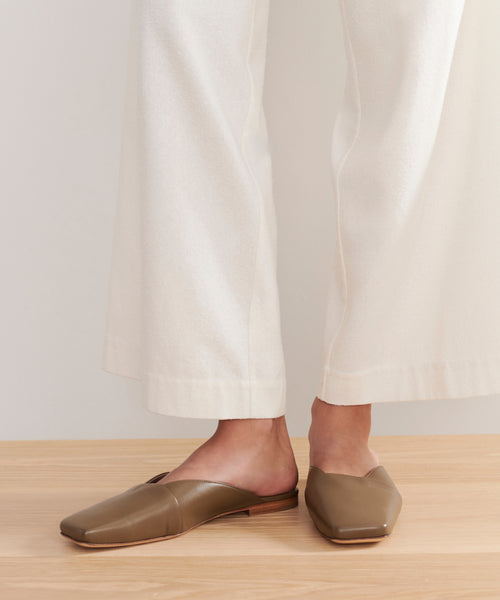 A person stands on a light wooden floor wearing cream wide-leg pants and the Mia Mule—light brown Italian leather, square-toe slip-ons with a minimalist design and open back.