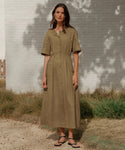 A woman with long brown hair stands on a gravel path by a white brick wall, wearing an olive green, short-sleeved button-down midi dress and Leather Mila Sandals. She has a relaxed expression.