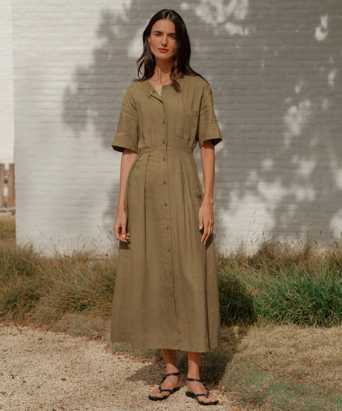 A woman with long brown hair stands on a gravel path by a white brick wall, wearing an olive green, short-sleeved button-down midi dress and Leather Mila Sandals. She has a relaxed expression.