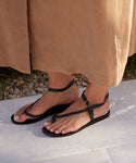 A person stands on a light surface, wearing a tan skirt and the Leather Mila Sandal—black braided leather sandals with ankle straps. Pebbles and greenery appear in the background.