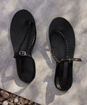 A pair of black Leather Mila Sandals with flat braided leather straps and buckles rests on a concrete surface with small pebbles in the corner, casting soft shadows.