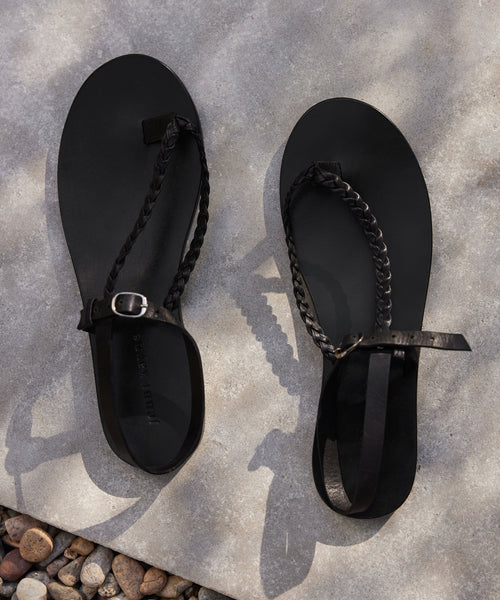 A pair of black Leather Mila Sandals with flat braided leather straps and buckles rests on a concrete surface with small pebbles in the corner, casting soft shadows.