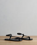 The Leather Mila Sandal, a pair of black braided leather sandals with ankle straps, is displayed side by side on a wooden surface against a plain light gray background.